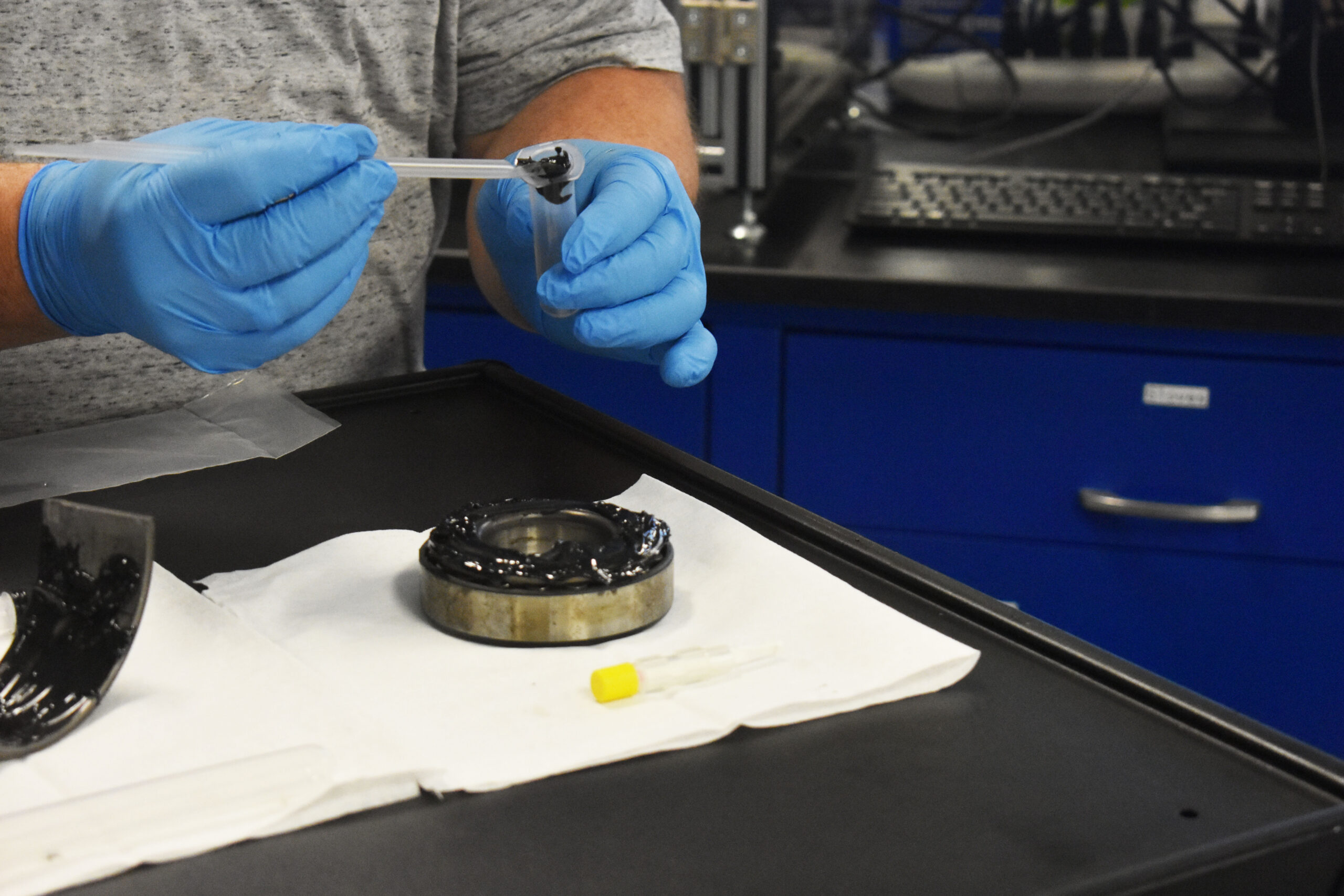 Technician in blue nitrile gloves collecting a grease sample from a bearing using a syringe sampling tool in a laboratory setting